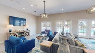 Located in Ponte Vedra Beach, Florida, this single-story clubhouse is designed with a vibrant coastal aesthetic that sets a welcoming tone throughout. The main gathering area is generously proportioned, featuring light wood-look flooring, recessed lighting, and statement geometric pendant fixtures that anchor the room with modern style. Comfortable seating arrangements — including plush blue velvet sofas, a tufted gray sectional, and accent chairs in rich teal and cobalt tones — are centered around natural live-edge wood coffee tables, reinforcing the organic, beach-inspired theme. A large wall-mounted flat-screen TV and a barn-wood media console complete the lounge area. Multiple sets of white-framed French doors flood the space with natural light and provide direct views of and access to the outdoor area, likely a pool. A decorative console table adorned with teal vases and a colorful framed mirror adds an elegant accent corner. The kitchen area features white cabinetry, granite countertops, stainless steel appliances including a refrigerator and microwave, and a breakfast bar-style counter that opens to the main gathering space. Coastal artwork throughout ties the design together, creating a cohesive, relaxed atmosphere ideal for community gatherings.