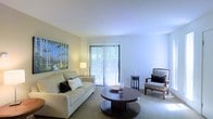 This single-floor residence offers a clean, well-appointed layout designed for comfortable everyday living. The living room serves as the heart of the space, showcasing a bright and airy atmosphere enhanced by large sliding glass doors that flood the room with natural light and provide direct access to an outdoor area. Vertical blinds offer adjustable light control while maintaining a sleek, uncluttered look. The room features soft, neutral-toned carpeting that complements the light walls and smooth white ceiling, creating a cohesive and calming aesthetic throughout. A comfortable sofa paired with a classic accent chair and a round wooden coffee table gives the living area a welcoming and functional arrangement. Decorative touches, including a nature-inspired wall painting, add a subtle artistic character to the space. The overall ambiance is serene and understated, with a warm, residential feel supported by thoughtful lighting from a standing lamp. The neutral palette and natural light make the space feel open and versatile, well-suited for both relaxing and entertaining.