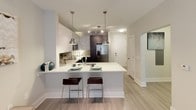 This single-floor residence showcases a contemporary design aesthetic with a carefully curated two-tone color palette. The kitchen is a standout feature, outfitted with high-gloss white upper cabinets contrasted by dark espresso lower cabinetry, all complemented by crisp white countertops and a neutral tile backsplash. Stainless steel appliances, including a French door refrigerator, built-in microwave, and range, reinforce the modern character of the space. A peninsula with pendant lighting and bar seating provides an efficient casual dining option while maintaining an open connection to the living area. Light wood-style flooring runs throughout the main living spaces, adding warmth to the clean-lined design. The living area features neutral walls that offer a versatile backdrop. The bedroom is carpeted for comfort and includes a ceiling fan, natural light from windows with blinds, and ample wall space. The bathroom is finished with a dark-cabinet vanity topped with a white countertop, a large mirror, and a tub-shower combination with white tile surround. Throughout, the design balances functionality with a polished, modern sensibility.