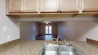 This single-floor space offers a practical and cohesive open-concept layout that connects the kitchen directly to the living area, creating a seamless flow throughout. The kitchen is outfitted with light natural wood cabinetry providing generous overhead storage, complemented by granite-patterned countertops that add texture and durability to the workspace. A stainless steel double-basin sink with a brushed nickel faucet serves as a functional centerpiece. The pass-through design between the kitchen and living area enhances the sense of openness, allowing easy visual connection between both spaces. The living area features dark hardwood flooring that contrasts attractively with the neutral, warm-toned walls. A ceiling fan provides both lighting and air circulation. The room is anchored by a set of French doors at the far end, which allow natural light to filter into the space while offering potential access to an outdoor area. The overall ambiance is clean, functional, and understated, with a neutral color palette that provides a versatile backdrop suitable for a variety of interior styles.