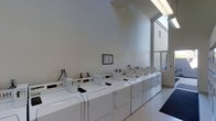 This shared laundry facility is designed with practicality and efficiency in mind, offering residents a clean and well-maintained space for their laundry needs. The room features a row of commercial-grade top-loading washing machines paired with matching dryers, all neatly aligned to maximize the use of the available space. The machines are equipped with digital control panels, making them straightforward to operate.

The facility benefits from soaring ceilings that contribute to an open, airy atmosphere, preventing the space from feeling cramped despite its focused purpose. A large window along one wall allows ample natural light to fill the room, enhancing visibility and creating a bright, welcoming environment. The neutral white walls and light-toned flooring reinforce the clean aesthetic throughout.

An informational notice board is mounted on the wall, providing users with relevant guidelines and instructions for operating the machines. A utility connection panel is also accessible along the wall, ensuring all equipment remains properly serviced. The entrance features a practical floor mat, helping to maintain cleanliness within the space. Overall, this laundry facility is thoughtfully arranged to provide a functional, comfortable, and efficient experience for all users.