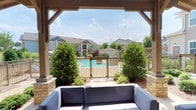 This charming gazebo serves as a dedicated outdoor lounge space within a residential community, designed to provide both shade and a scenic vantage point. The structure features sturdy corner posts with stone bases and a wooden overhead framework, creating an open yet sheltered environment ideal for relaxation.

Inside, plush sectional seating with cushioned upholstery invites residents to unwind in comfort. The open design of the gazebo allows for excellent airflow and unobstructed sightlines to the surrounding community amenities.

Directly ahead, a gated community swimming pool takes center stage, framed by neatly manicured shrubs and ornamental trees that add lush greenery to the setting. The pool area is well-maintained and enclosed by a decorative iron fence, with lounge areas visible beyond the gate. The backdrop features the community's residential buildings set against a bright, open sky with mature trees lining the perimeter.

The overall ambiance is bright, open, and inviting, making this gazebo an ideal spot for casual socializing or quiet enjoyment of the outdoors. The thoughtful landscaping and proximity to the pool create a resort-like atmosphere that enhances the outdoor living experience for the community.