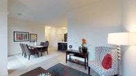 This single-floor residence is designed around an open-concept layout that effortlessly connects the living, dining, and kitchen areas into one cohesive and functional living space. The interior is finished with light hardwood-style flooring throughout, contributing to a warm yet contemporary feel that is consistent across all areas.

The living area provides a comfortable and well-lit space, featuring crisp white walls and smooth ceilings with recessed lighting that ensures even, ambient illumination. A patterned area rug anchors the seating arrangement, adding a touch of texture and color to the neutral palette.

Adjacent to the living space, the dining area accommodates a full dining table with seating for multiple guests, set beneath natural and artificial lighting that creates an inviting atmosphere for everyday meals or entertaining.

The kitchen is sleekly designed with dark cabinetry contrasting against light countertops and stainless steel appliances. A breakfast bar with bar stools provides casual seating and serves as a natural transition between the kitchen and the rest of the open living space. Pendant lighting above the bar adds a refined, modern detail.

Overall, the space presents a clean, minimalist aesthetic with thoughtful design choices that balance functionality and style, making it a comfortable and visually appealing environment.
