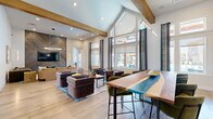 This thoughtfully designed single-floor space offers an impressive blend of architectural drama and contemporary style. Soaring vaulted ceilings with exposed wood beams define the open layout, while expansive floor-to-ceiling windows and glass gable features flood the interiors with abundant natural light and provide views of the surrounding exterior. Light-toned hardwood flooring flows seamlessly throughout, unifying the distinct functional areas.

The living room is a standout, featuring multiple seating zones that cater to both relaxed lounging and social gathering. A striking wood-paneled accent wall with a mounted television anchors one lounge area, while a bold marble-effect feature wall with another mounted display and a billiards table create an entertainment-focused zone. Curated furnishings—including a live-edge wooden bar counter with teal resin inlay, velvet lounge chairs, and a leather sofa—add warmth and texture. A large circular pendant light serves as an elegant focal point overhead.

The office area is bright and functional, equipped with a built-in desk workstation positioned beneath generous windows that offer ample natural light. Additional workspace with seating for multiple users makes it well-suited for productivity. Geometric wall paneling and exposed brick accents add visual interest throughout, contributing to the space's cohesive yet eclectic design aesthetic.