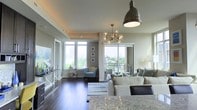 This thoughtfully designed single-floor residence offers a seamless open-concept layout that connects the living, dining, and kitchen areas into one cohesive, light-filled space. Dark hardwood flooring runs throughout, providing a rich contrast against the soft, neutral-toned walls and contemporary furnishings. The kitchen features granite countertops and flows effortlessly into the dining and living areas, making the space ideal for both entertaining and everyday living. Statement lighting anchors the interior, including a sleek industrial pendant over the kitchen and a multi-bulb sputnik chandelier that adds a modern artistic touch to the living area. The living room is generously proportioned, centered around a comfortable sectional sofa, and benefits from an abundance of natural light streaming in through expansive windows and sliding glass doors. These doors open onto a private balcony, where sweeping views of the surrounding cityscape and lush tree canopy can be enjoyed. A cozy reading nook with a cushioned armchair is nestled near the windows, offering a peaceful retreat within the home. The overall ambiance is modern and sophisticated, with carefully curated décor that balances style and comfort throughout.