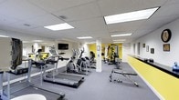 This single-floor fitness center is thoughtfully designed to accommodate a wide range of workout routines. The space is outfitted with a strong selection of cardio equipment, including treadmills and elliptical trainers, all positioned on protective floor mats for safety and comfort. A multi-station strength training machine anchors one side of the room, complemented by free weights and additional resistance equipment for a complete strength training experience. The flooring throughout is a durable grey carpet, well-suited for a high-traffic exercise environment. Bright, recessed panel lighting in the drop ceiling ensures the space is evenly and amply illuminated. Bold yellow accent walls inject energy into the room, and motivational quotes are painted directly onto the walls, reinforcing an encouraging atmosphere. A wall-mounted flat-screen television provides entertainment during workouts. Structural columns are cleanly finished and integrated into the layout without disrupting the flow of the space. The overall design is functional and practical, balancing an open, airy feel with a well-organized arrangement of equipment to maximize usability for a variety of fitness levels and goals.
