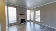 This single-story, one-bedroom, one-bathroom residence offers a well-appointed living space designed with both comfort and style in mind. The living area serves as the focal point of the unit, showcasing warm hardwood-style flooring that runs throughout and reflects the abundant natural light streaming in from multiple large windows. Crown molding frames the ceiling, lending a refined architectural detail that elevates the overall aesthetic.

A classic white fireplace with a clean mantel serves as a striking centerpiece along one wall, adding both visual interest and a cozy ambiance to the room. Neutral warm-gray wall tones complement the light-toned flooring, creating a cohesive and inviting palette throughout the space.

Large windows on multiple walls ensure the living area remains bright and airy, while a glass-paned door provides direct access to a private balcony, offering outdoor views and additional ventilation. The balcony extends the living experience beyond the interior, providing a dedicated outdoor retreat.

Overall, the space combines functional design with thoughtful architectural details, making it a comfortable and visually appealing single-floor residence.