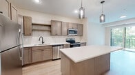 Located at 6701 W 78th St in Bloomington, MN, this one-bedroom, one-bathroom unit offers a well-designed single-floor layout with a contemporary aesthetic and quality finishes throughout. The kitchen is a standout feature, showcasing warm taupe shaker-style cabinetry, a subway tile backsplash, crisp white quartz countertops, and a full suite of stainless steel appliances including a refrigerator, range, microwave, and dishwasher. A generous kitchen island with pendant lighting above provides additional prep space and a natural gathering point. Light hardwood-style flooring flows through the main living and kitchen areas, lending warmth and continuity. The bedroom is a comfortable retreat, finished with soft carpeting and featuring a large window and sliding glass door that open to a private balcony, offering pleasant tree-lined views and abundant natural light. A dedicated walk-in closet provides practical storage. The bathroom is finished with a wood-toned vanity, white quartz countertop, bold black hardware and mirror frame, and hardwood-style flooring, creating a clean, modern look. The overall ambiance is bright and contemporary, with neutral tones and natural greenery views contributing to a calm, welcoming atmosphere throughout.