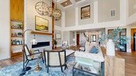 This thoughtfully designed single-floor amenity space combines dramatic architectural elements with carefully curated interiors throughout several distinct functional areas. The centerpiece is a grand living lounge showcasing soaring double-height ceilings with coffered detailing, a floor-to-ceiling stacked stone fireplace framed by warm wood accents, and a striking pair of oversized orb chandeliers that anchor the space. Large clerestory windows draw natural light deep into the room, complementing the rich wood-look flooring that flows throughout. Comfortable seating arrangements, including tufted sofas, cane-back chairs, and glass-topped coffee tables over a blue patterned rug, create an inviting atmosphere for gathering.

Adjacent to the main lounge, a kitchen and bar area features warm wood cabinetry, a herringbone tile backsplash, stainless steel appliances, a butcher block island with bar seating, and glass pendant lighting overhead. A bright, open entry corridor with soaring windows and a staircase leads residents through the space seamlessly. A dedicated dining room with teal accent walls, wood-framed art, and natural light from multiple windows provides a relaxed setting for casual meals, while additional lounge seating near expansive windows offers pleasant outdoor courtyard views.