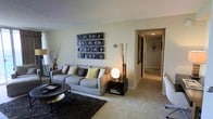 This single-floor residence offers a thoughtfully arranged living space with a warm and contemporary aesthetic. The living room serves as the centerpiece, featuring plush wall-to-wall carpeting that adds comfort underfoot and a cozy atmosphere throughout. A generously sized gray sofa anchors the seating area, complemented by an assortment of accent pillows in neutral and earthy tones. A dark area rug grounds the space beneath a sleek, round coffee table, adding contrast and definition to the room. A striking grid-style wall art installation in dark tones serves as a bold focal point above the sofa, while decorative branch arrangements and a spherical accent lamp contribute to the room's modern sensibility. Neutral, light-toned walls keep the space feeling open and airy, and recessed lighting provides a clean, unobtrusive source of illumination. A hallway extends from the living area, hinting at additional rooms beyond, with doors visible along its length and consistent carpeting carried throughout. The overall ambiance is calm and inviting, with a balanced mix of functional furnishings and decorative touches that create a polished, livable environment.
