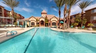 This expansive outdoor amenity space is designed for relaxation and entertainment, centered around a large, sparkling swimming pool with striking turquoise water. The pool area is framed by tall palm trees and a Mediterranean-inspired clubhouse building with a tile roof, arched windows, and warm terracotta and stucco exterior finishes that lend the space a resort-like character.

Adjacent to the main pool is a circular hot tub, surrounded by a generous deck lined with cushioned lounge chairs and framed by an elegant curved pergola structure. A dedicated shaded cabana zone features a slatted canopy overhead, teal-colored chaise lounges, privacy curtains, and small side tables — ideal for extended outdoor relaxation.

The covered lounge pavilion is equipped with an outdoor fireplace finished in coordinating terracotta-toned tile, wall-mounted television, and comfortable seating arranged to encourage social gathering. Natural light filters through open clerestory windows, keeping the space bright and airy even when shaded.

A fully outfitted outdoor kitchen rounds out the amenities, featuring a large built-in stainless steel grill, ample granite counter space, a utility sink, and durable anti-fatigue flooring — all set beneath a covered structure with decorative iron railings and views of the surrounding landscaped courtyard. Together, these areas create a cohesive, well-appointed outdoor living destination.