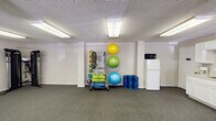 Located at 115 Holiday Park Rd in Hillsborough, NC, this single-floor fitness center offers a well-equipped and functional workout facility. The main exercise area is spacious and open, with carpeted flooring, textured ceilings, and bright fluorescent lighting throughout. Large windows along one wall allow natural light to filter in, creating an inviting atmosphere. The space accommodates a variety of workout styles, featuring cardio equipment including an elliptical machine and treadmill, as well as an Inspire functional trainer cable machine for strength training. A wall-mounted television is positioned for easy viewing during workouts. A dedicated equipment storage area houses an organized rack of medicine balls, resistance bands, and exercise mats, alongside a collection of stability balls in multiple sizes and rolled yoga mats. A compact kitchenette area provides convenience with white cabinetry, a countertop sink, a coffee maker, and a mini refrigerator with microwave, making it easy for users to stay refreshed. A glass-paned exterior door offers direct access to the outdoors. The overall layout is clean, practical, and well-maintained, providing a comfortable and complete fitness environment suited for a range of exercise routines.
