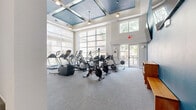 Located at 3333 Weslayan St in Houston, Texas, this fitness center offers a generously sized, single-floor layout divided into two distinct exercise areas connected by a hallway. The cardio room is an airy, light-filled space defined by soaring ceilings with striking teal-painted coffered accents and floor-to-ceiling windows that flood the room with natural light and offer views of the surrounding greenery. This area is equipped with a full lineup of Matrix treadmills, ellipticals, stationary bikes, and a rowing machine, complemented by a wall-mounted flat-screen TV and pendant lighting. A textured stone-clad reception counter anchors the transitional hallway area, which also features a mounted TV and seating, serving as a welcoming entry point between the two zones. The strength training room features a rich teal ceiling with recessed lighting and rubber flooring for safety and durability. It is outfitted with a comprehensive free weight selection, weight benches, a cable machine, a multi-station strength system, and full-length mirrors that enhance the sense of depth. Built-in shelving provides organized storage for smaller equipment. Together, both rooms deliver a complete fitness experience in a modern, well-designed environment.