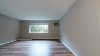 This single-floor unit is thoughtfully laid out to maximize functionality across its living spaces. Throughout the unit, wood-look laminate flooring in warm and cool tones creates a cohesive aesthetic, complemented by neutral grey walls and white baseboards.

The living area is generously proportioned, featuring a large window that frames outdoor greenery and allows ample natural light to fill the room. A wall-mounted air conditioning unit and baseboard heater ensure year-round climate comfort. The open sightline from the entryway into the living space creates a welcoming, connected feel.

The galley-style kitchen is efficiently designed with rich, dark wood cabinetry lining both sides, granite-look countertops, and stainless steel appliances including a refrigerator, range, and dishwasher. Overhead cabinetry provides substantial storage throughout.

The bedrooms are well-sized, each featuring windows with outdoor views, baseboard heating, and wall-unit air conditioning, ensuring individual climate control. The light flooring and neutral walls contribute to a clean, versatile atmosphere.

The bathroom is finished with white subway-style wall tiles surrounding a full soaking tub, wood-look flooring, and classic white fixtures, offering a tidy and functional space.
