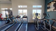 Located on Outlet Blvd in Fort Worth, TX, this single-level fitness center is designed to support a comprehensive workout experience. The space features high ceilings and an open, airy layout that contributes to a motivating atmosphere throughout.

The cardio zone is lined with large, multi-pane windows that flood the area with natural light while offering views of the surrounding outdoor pool area and landscape. This section houses multiple treadmills, elliptical machines, and upright and recumbent stationary bikes, all positioned to take advantage of the natural light and outdoor scenery.

The strength training area is equally well-appointed, featuring a variety of cable and weight machines, free weights, and a large wall-mounted mirror that enhances the sense of space. A standout feature of this zone is a Freedom Climber rotating climbing wall, providing a unique and engaging full-body training option. A large display screen is also present, offering entertainment or guided workout options.

Additional equipment includes a stair climber positioned near the exit corridor, which connects to an adjacent room with glass-panel doors. The flooring throughout is durable rubber gym matting, suitable for high-impact activity. The clean, white walls and recessed lighting further contribute to a bright, professional environment built for fitness.