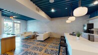 Located at 3110 Grand Ave in Pinellas Park, Florida, this single-floor commercial space is a thoughtfully designed coffee lounge and coworking destination. The interior immediately impresses with its striking teal slatted ceiling, which anchors the space with a bold architectural statement and contrasts elegantly against crisp white walls clad in large-format tile.

The flooring throughout is a standout feature — a distinctive pattern of terrazzo insets framed by warm wood-toned planks creates a dynamic visual rhythm underfoot. Seating is varied and purposeful: plush cream lounge sofas with integrated side tables offer a relaxed environment, while a long communal work table positioned near floor-to-ceiling windows provides a collaborative workspace bathed in natural light and views of the surrounding Florida landscape.

A built-in banquette with woven accent chairs lines one wall, accompanied by curated artwork, making it ideal for casual meetings or quiet work sessions. The coffee bar area is fully equipped with a white quartz island, professional espresso and brewing equipment, a beverage cooler, and globe pendant lighting that adds warmth to the service zone. The overall ambiance is modern and inviting, balancing functionality with a refined aesthetic suited for both productivity and leisure.