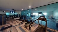 This contemporary fitness center is thoughtfully designed to support a full range of workout activities within a single, open-plan space. The flooring throughout features a striking herringbone-patterned hardwood in light and mid-tone wood tones accented with grey inlays, lending the space a polished, upscale aesthetic.

The cardio zone is well-stocked with a lineup of treadmills, elliptical trainers, and recumbent bikes, each equipped with individual display screens for interactive workout tracking. Adjacent to this, a dedicated strength training area offers a robust selection of dumbbells arranged on tiered racks, weight benches, and multi-function cable machines from Matrix and other commercial-grade brands.

A defining architectural feature is the full-height teal-tinted glass wall partition, which adds depth, transparency, and a sleek modern character to the interior. The space is lit by a combination of recessed ceiling lights and a suspended linear pendant fixture, ensuring bright, even illumination throughout. A ceiling fan adds to the comfort of the workout environment.

Wall-mounted flat-screen televisions are positioned for easy viewing across the floor. Mirrored walls in the free weights section expand the sense of space and aid in form monitoring. A clean, neutral color palette of soft grey and white walls completes the refined and functional atmosphere of this fitness facility.