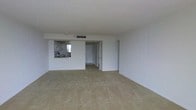This single-floor space presents an open-concept living area characterized by clean white walls and a neutral-toned carpet that extends throughout the main room, creating a blank canvas well-suited for a variety of interior styles. The layout is straightforward and functional, with the living area flowing seamlessly toward the entryway and additional rooms beyond.

A pass-through kitchen window connects the kitchen to the main living space, allowing natural light from the kitchen's exterior window to filter into the interior. The kitchen itself is partially visible through the pass-through, hinting at a compact but practical cooking area. The pass-through design also supports an open, communicative atmosphere between spaces.

The living area features a textured ceiling with centrally placed HVAC venting, suggesting a climate-controlled environment. Several doorways are visible from the main room, indicating access to additional private spaces such as bedrooms or storage areas. The overall ambiance is understated and utilitarian, with white walls and minimal architectural detailing keeping the focus on the spatial layout. The unit offers a functional foundation suitable for comfortable everyday living.