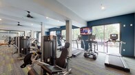 Located at 891 Ingleside Ave in Columbus, Ohio, this well-appointed fitness center offers a comprehensive single-floor layout designed to support a wide range of workout routines. The space is defined by its modern aesthetic, featuring a bold navy and neutral color palette, recessed lighting, and multi-toned carpet tile flooring throughout.

The cardio zone is lined with True-brand treadmills, ellipticals, and cycling machines positioned along floor-to-ceiling glass walls that flood the area with natural light and provide views of the surrounding greenery and outdoor terrace. Wall-mounted flat-screen televisions are strategically placed throughout for convenient viewing during workouts.

A dedicated strength training area houses a selection of selectorized machines and cable equipment, complemented by a free-weight station stocked with dumbbells, medicine balls, and stability balls. Full-length mirrored walls in the studio area expand the sense of space while supporting stretching, yoga, or group fitness activities.

Additional amenities include a refreshment station with cabinetry, ceiling fans for comfort, and direct access to an outdoor area. The open floor plan allows for easy movement between zones, and the generous spatial layout accommodates multiple users simultaneously without feeling crowded.