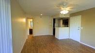 This single-floor space features an open-concept design that seamlessly connects the living area to the kitchen. The living area is finished with warm wood-style flooring that extends throughout, complemented by neutral-toned walls and white baseboards that create a clean, cohesive aesthetic. A ceiling fan with a built-in light fixture is centrally positioned, providing both air circulation and ambient lighting alongside recessed ceiling lights. The textured ceiling adds a subtle dimension to the space.

The kitchen is defined by a raised breakfast bar with granite-style countertops, offering a natural division between the living and cooking areas. Dark cabinetry lines the kitchen walls, providing a striking contrast against the lighter surroundings. A refrigerator and additional appliances are visible within the kitchen space.

Built-in open shelving on one wall offers practical display or storage options in the living area. A hallway visible from the main space leads to additional rooms, including what appears to be a bathroom with matching cabinetry. A closet door is also present near the kitchen, adding to the functional storage solutions within this well-appointed space.
