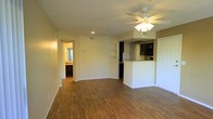This single-floor space features an open-concept design that seamlessly connects the living area to the kitchen. The living area is finished with warm wood-style flooring that extends throughout, complemented by neutral-toned walls and white baseboards that create a clean, cohesive aesthetic. A ceiling fan with a built-in light fixture is centrally positioned, providing both air circulation and ambient lighting alongside recessed ceiling lights. The textured ceiling adds a subtle dimension to the space.

The kitchen is defined by a raised breakfast bar with granite-style countertops, offering a natural division between the living and cooking areas. Dark cabinetry lines the kitchen walls, providing a striking contrast against the lighter surroundings. A refrigerator and additional appliances are visible within the kitchen space.

Built-in open shelving on one wall offers practical display or storage options in the living area. A hallway visible from the main space leads to additional rooms, including what appears to be a bathroom with matching cabinetry. A closet door is also present near the kitchen, adding to the functional storage solutions within this well-appointed space.