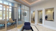 This single-floor business center is thoughtfully designed to support a variety of professional needs in a polished and welcoming environment. The main area is anchored by individual workstations with ergonomic seating, providing comfortable spaces for focused, independent work. Light wood-look tile flooring runs throughout, complemented by neutral walls and tray ceilings with subtle ambient lighting that create a bright, professional atmosphere. A glass-walled conference room offers excellent visibility and natural light, furnished with a full complement of rolling chairs and a large table—ideal for collaborative meetings or presentations. Patterned drapery and floor-to-ceiling windows add an elegant touch while maintaining a connection to the outdoors. Multiple private offices or study rooms, each distinguished by decorative honeycomb-patterned accent walls and individual desks, provide quiet spaces for focused work or private calls. A dedicated refreshment station, set against a striking textured stone accent wall, includes a coffee setup and storage cabinetry. Wall-mounted televisions enhance the functionality of the common areas. An adjacent recreation space with a billiards table is visible from the main area, rounding out this comprehensive and versatile business center.