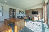 Located in Atlanta's Bolton Drive corridor, this single-floor amenity lounge blends industrial character with contemporary design. Exposed brick columns and accent walls provide a warm, textural contrast against smooth neutral-toned walls and dark slate-style carpeting throughout the space.

Natural light pours in through expansive floor-to-ceiling windows framed in sleek black steel, offering generous views of the surrounding courtyard, pool area, and neighboring architecture. The open layout is divided into distinct functional zones that flow seamlessly into one another.

A collaborative work area features a long built-in wooden desk with ample surface space, paired with modern swivel chairs—ideal for focused work or casual meetings. Adjacent to this, a lounge zone offers leather armchairs with plaid accent pillows, centered around a low coffee table, creating an inviting spot for relaxed conversation or informal gatherings.

A solid wood communal table with matching bench seating anchors a central area, well-suited for group work or social dining. Curated wall art, including an abstract canvas and a gallery of framed prints, adds a polished, thoughtful aesthetic throughout. Steel-framed glass doors connect the space to adjacent areas, enhancing its open, connected feel. The overall ambiance is sophisticated yet approachable, balancing productivity and comfort.
