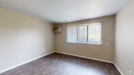 This single-floor unit offers a practical and cohesive layout connecting the main living area, kitchen, bedroom, and bathroom. The living spaces are finished with durable wood-look flooring in a warm, medium-toned brown, complemented by neutral beige walls and textured ceilings throughout. Wall-mounted air conditioning units provide climate control in the main rooms, and vertical blinds on the windows offer both privacy and natural light, with outdoor greenery visible through the glass.

The kitchen is well-equipped with white shaker-style cabinetry extending to the upper walls for ample storage, granite-patterned countertops, and a full suite of stainless steel appliances including an electric range, dishwasher, and refrigerator. A peninsula counter provides additional workspace and separates the kitchen from the adjoining living area, which is further enhanced by a ceiling fan overhead.

The bedroom features plush carpeting and a window fitted with vertical blinds, creating a comfortable and quiet retreat. The bathroom includes a full tub and shower combination with white tile surround, a toilet, and a vanity with a granite-style countertop, all set on wood-look flooring consistent with the rest of the unit.