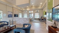 Located on Lake Nona Boulevard in Orlando, Florida, this single-floor community clubhouse offers a thoughtfully designed gathering space with a contemporary coastal aesthetic. The open-plan layout is anchored by soaring ceilings and an abundance of natural light streaming through expansive floor-to-ceiling windows that frame views of the surrounding Florida landscape, including lush palm trees.

The main lounge area features a striking natural stone accent wall with a wall-mounted flat-screen television, complemented by plush neutral sofas accented with green and teal throw pillows. Wide-format tile flooring flows throughout, giving the space an airy, cohesive feel. A set of glazed double doors opens directly to an outdoor area, seamlessly connecting the interior to the exterior amenities.

A dedicated dining and social zone showcases a live-edge communal table paired with contemporary gray chairs, set against a vibrant green mosaic tile feature wall with an additional mounted display. A kitchenette and refreshment station serve this area, making it well-suited for community gatherings.

A bright sitting lounge with double-height windows offers intimate seating nooks bathed in sunlight, ideal for casual conversation or quiet relaxation. Bold color-block drapery adds visual interest throughout. Decorative art pieces, a chandelier, and carefully curated furnishings complete the polished, welcoming ambiance of this versatile community space.