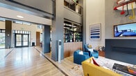 This contemporary two-story amenity facility is thoughtfully designed to offer a diverse range of social, recreational, and professional spaces. The grand lobby makes an immediate impression with its soaring double-height ceilings, bold structural columns, light hardwood flooring, and a curated seating area featuring vibrant teal and yellow upholstered chairs arranged around a central fireplace. A decorative open shelving unit with live plants adds a natural element, while a striking wall-mounted art piece anchors the space above the fireplace.

The second floor, accessible via a staircase with sleek metal railings, overlooks the lobby and features warm hardwood flooring with views down to the main level. On the ground floor, a reception desk with a dark countertop and wood paneling faces glass-enclosed conference and meeting rooms outfitted with modern furnishings and neon accent signage.

A dedicated game room offers a shuffleboard table, a classic arcade cabinet, and exposed brick walls adorned with large-format electric guitar photography. Lounge seating and floor-to-ceiling windows provide outdoor views of the surrounding courtyard. A separate media lounge features a built-in dual-screen television display with a wood surround, carpeted flooring, and comfortable seating. Throughout, the space blends industrial materials with warm accents and curated artwork for a cohesive, energetic atmosphere.
