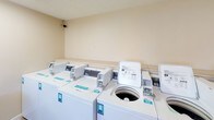 The Georgetown South laundry facility is a well-organized, single-floor communal space designed to meet the everyday needs of its residents. The room is laid out efficiently, housing a generous row of top-loading washing machines and stacked dryer units lining the walls, each equipped with a coinless payment system for added convenience. The machines are numbered and clearly labeled, making it easy to identify and manage laundry loads.

Natural light filters in through window blinds, brightening the neutral, cream-toned walls and tile flooring that give the space a clean, functional aesthetic. Overhead fluorescent lighting ensures the room remains well-lit throughout the day. A dedicated folding area features multiple foldable tables, providing ample surface space for sorting and folding laundry. A built-in counter with cabinet storage below offers additional workspace near a window overlooking the parking area.

A vending machine is conveniently located near the entrance, and a community bulletin board keeps residents informed of local notices and updates. Security cameras are positioned throughout the space, ensuring a safe environment. A waste bin is also provided for resident use. The overall layout is straightforward and practical, making this laundry facility a reliable and accessible amenity for the Georgetown South community.