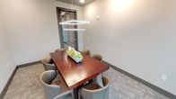 This single-floor professional office suite is thoughtfully designed to support a focused and productive work environment. The centerpiece of the space is a private conference room finished with neutral tones and patterned carpet tiles that add subtle texture underfoot. A rich, warm-toned rectangular conference table seats up to six in contemporary upholstered chairs, creating an inviting setting for meetings and collaboration. Overhead, a striking square pendant light fixture serves as a design focal point while providing ample illumination. Two large-scale abstract artworks in muted golds and grays adorn the wall, lending a refined, curated aesthetic to the room. A glass-paneled door connects the conference room to the adjacent office area, maintaining visual openness while offering acoustic privacy. The adjoining office area benefits from generous floor-to-ceiling windows framed in dark metal, flooding the space with natural light and offering views of the surrounding landscaped exterior. Recessed ceiling lights throughout ensure consistent, even illumination across all areas. The overall design balances professionalism with comfort, featuring clean architectural lines, neutral wall finishes, and carefully selected furnishings that create a cohesive and functional workspace.