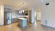 This single-floor unit showcases a contemporary open-concept layout with a strong emphasis on the kitchen as its central feature. The kitchen is outfitted with flat-front gray cabinetry extending to the ceiling, offering generous storage capacity. Crisp white quartz countertops contrast cleanly against the gray tones, while a white subway tile backsplash adds subtle texture. A full suite of stainless steel appliances includes a French door refrigerator, electric range, built-in microwave, and dishwasher. A freestanding center island with a matching white countertop provides additional prep space and casual seating potential. Pendant lighting on adjustable hanging rods illuminates the island, complemented by a track lighting system overhead. Light-toned hardwood-style flooring runs continuously throughout, enhancing the sense of openness and connecting the kitchen seamlessly to the living area. The living space features a ceiling fan and a raised ceiling detail, adding architectural dimension. A frosted glass interior door introduces a modern design element while maintaining a bright, airy feel. Neutral white walls and consistent finishes throughout contribute to a clean, cohesive aesthetic well-suited for modern living.