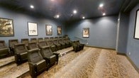 This dedicated private theater room delivers an immersive cinematic experience with a thoughtfully designed layout and premium finishes throughout. The room features tiered, stadium-style seating arranged across two elevated levels, ensuring unobstructed sightlines from every seat. The plush, dark leather recliner-style chairs are modeled after classic cinema seating, complete with armrests and individual seat dividers, providing both comfort and an authentic movie-house atmosphere.

The ceiling is finished in a deep, dark tone that minimizes light reflection and enhances the viewing environment, while recessed ceiling lighting offers ambient illumination when needed. A ceiling-mounted projector is centrally positioned to deliver a high-quality projection onto the screen. The walls are painted in a rich slate blue, adding to the moody, cinematic ambiance of the room.

Classic framed movie posters adorn the walls, lending a nostalgic nod to Hollywood's golden era and reinforcing the space's dedicated entertainment purpose. The floor is covered in a richly patterned carpet that adds warmth and aids in sound absorption. A handrail along the stepped aisle ensures safe and easy navigation between seating tiers. Overall, this screening room is a well-appointed, purpose-built venue designed to replicate the full theater-going experience in a private setting.