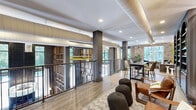 Located at 215 Reformation Pkwy in Canton, GA, this well-appointed hallway space delivers a refined communal atmosphere that blends industrial character with contemporary design. The single-floor layout is anchored by wood-look tile flooring that runs the full length of the space, complemented by bold dark structural columns that define zones without closing off the open feel.

An open metal railing system lines one side, offering views to the level below and inviting natural light from large windows that frame lush outdoor greenery. Exposed ductwork and recessed lighting contribute to the space's modern-industrial aesthetic, while original brick accent walls add warmth and texture throughout.

Distinct seating vignettes are thoughtfully arranged along the corridor — from tufted sofas and cushioned lounge chairs to sculptural ottomans and high-top dining sets — making the space as functional as it is visually engaging. A standout design feature is a backlit geometric wall panel with bold yellow cross-brace accents, paired with a set of dark arched built-in shelving units that add depth and sophistication. Curated artwork and lush greenery complete the inviting, gallery-like atmosphere, making this hallway a destination in its own right rather than simply a passageway.