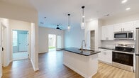 Located at 214 Kingsmeadow Cir in Belville, NC, this thoughtfully designed single-floor residence offers a clean, modern aesthetic throughout. The open-concept kitchen and living area serves as the heart of the home, anchored by a central island with dark granite countertops, a stainless steel sink, and elegant pendant lighting overhead. White shaker-style cabinetry, a subway tile backsplash, and a full suite of stainless steel appliances—including a refrigerator, range, and microwave—complete the kitchen. Light wood-look flooring flows seamlessly through the living and kitchen areas, while a ceiling fan and sliding glass door fill the living space with natural light and provide direct access to a private, fenced outdoor area with a lush green lawn. The bedroom features plush carpeting, a ceiling fan, natural light from a window with blinds, and access to a closet. The bathroom is finished with white cabinetry, dark granite countertops, a large vanity mirror, globe lighting, and wood-look flooring. A dedicated laundry room houses a full-size GE washer and dryer. The overall ambiance is bright, fresh, and move-in ready.