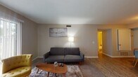 This single-floor residential unit offers a well-organized layout that balances comfort and functionality across several distinct living spaces. The living room serves as the central gathering area, featuring neutral beige walls, carpet flooring layered with a plush area rug, and a large sliding glass door fitted with vertical blinds that allows natural light to fill the room. A mounted flat-screen television and a mid-century style sofa and accent chair complete the space. Adjacent to the living room is a compact dining area with hardwood-style flooring, a round dining table, and access to a thermostat panel on the wall. The kitchen is galley-style, equipped with white cabinetry, granite-look countertops, white appliances including a refrigerator, electric range, dishwasher, and microwave, as well as an in-unit washer and dryer. The two bedrooms are comfortably sized with neutral tones, carpet flooring, ceiling-mounted ventilation, and ample natural light from windows with blinds. The full bathroom features a combined tub and shower with a curtain, a single-sink vanity with white cabinetry, and hardwood-style tile flooring. Throughout the unit, the consistent neutral palette and practical finishes create a clean, livable atmosphere.