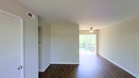 This single-floor space is thoughtfully laid out to maximize functionality and comfort. The living area serves as a central highlight, featuring rich, dark hardwood flooring that extends throughout the main living spaces, creating a warm and cohesive aesthetic. Neutral-toned walls complement the flooring and contribute to a clean, open atmosphere. A ceiling fan with a light fixture keeps the space comfortable and well-lit.

A standout feature of the living area is the large sliding glass door that opens to an outdoor balcony or patio, framing lush green tree views and drawing in generous amounts of natural light. Windows positioned throughout the unit further enhance the bright, airy feel of the interior.

The kitchen is positioned adjacent to the living space and offers a practical layout with standard appliances and cabinetry, providing efficient use of the available space. A bathroom is also included within the floor plan, visible in the overhead layout view.

The bedroom area is carpeted, offering a softer contrast to the hardwood found elsewhere. Overall, the space presents a functional and comfortable single-floor layout with a welcoming ambiance defined by natural light and consistent finishes.