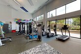 This fitness center is a thoughtfully designed, single-floor facility that combines functionality with an energizing aesthetic. The space is defined by its soaring double-height ceilings and expansive floor-to-ceiling windows along one full wall, flooding the interior with natural light and offering open views of the surrounding greenery and streetscape — creating an inviting, airy atmosphere ideal for exercise.

A striking large-scale mural in bold, multicolored abstract forms anchors one end of the space, adding a vibrant artistic focal point that elevates the energy of the room. The flooring transitions between patterned carpet tiles near the entry and open workout zones, providing both visual interest and practical zoning throughout.

The facility is comprehensively outfitted with a wide array of equipment to support diverse fitness routines. The cardio zone features multiple treadmills, elliptical machines, and a stair climber, all positioned to take advantage of the natural light and outdoor views. The strength and functional training area includes cable machines, a multi-use power rack, weight benches, a rowing machine, kettlebells, medicine balls, and suspension training cables. Full-length mirrors on one wall enhance the sense of space and support form-focused training.

The entrance is marked by a formal glass double-door entry with high ceilings, a Fitness Center information placard, and a patterned carpet that sets the tone for the polished, well-maintained environment within.