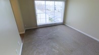 This single-floor space is thoughtfully laid out and offers a comfortable, functional environment. The living area is finished with wall-to-wall carpeting in a neutral tone, providing a warm and inviting foundation that complements the soft, muted wall colors throughout. A standout feature of the main living space is the expansive sliding glass door fitted with vertical blinds, which serves as the primary source of natural light. This large glazed opening not only brightens the interior but also frames views of the surrounding outdoor environment, including glimpses of neighboring structures and greenery. The transition between flooring materials — carpet meeting what appears to be a hard surface — adds a subtle delineation between areas of the unit. White baseboards provide a clean, crisp contrast to the neutral walls and flooring. The overall ambiance is modest and practical, offering a blank canvas well-suited for personalization. The space benefits from its elevated vantage point, as suggested by the outward views visible through the sliding door, contributing to a sense of openness despite the compact footprint.