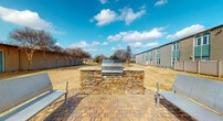 This community amenity space includes two distinct areas designed for resident convenience and outdoor enjoyment. The grill area features a built-in barbecue station constructed with stacked natural stone, topped with a stainless steel grill. The paved brick patio surrounding the station provides a comfortable setting, complemented by metal seating chairs on either side. Open skies and views of the surrounding grounds make this an inviting outdoor gathering spot.

Adjacent to the grill area is a well-equipped laundry facility, accessible through a paneled entry door with a sidelight window offering views of the outdoor patio. The laundry room features light wood-look flooring and neutral-toned walls that create a clean, functional environment. Multiple top-load and front-load washers are arranged alongside a full row of stacked dryers lining the back wall, providing ample capacity for residents. A ceiling fan ensures air circulation throughout the space, while windows allow natural light to filter in. A folding table is conveniently positioned along the wall, offering a practical surface for laundry tasks. The layout is open and efficiently organized for ease of use.