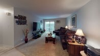 This single-floor residence at 640 S York Rd in Bensenville, Illinois offers a well-organized layout with distinct living spaces throughout. The living room is generously sized, finished with plush carpeting and neutral-toned walls. A large sliding glass door floods the space with natural light and provides direct access to an outdoor area, making it a bright and welcoming centerpiece of the home. The kitchen features white cabinetry, a peninsula countertop with granite-style laminate surfaces, and white appliances including a range and refrigerator. It connects seamlessly to a dedicated dining area, which includes a ceiling fan and room for a round dining table with a corner display shelf. The primary bedroom is carpeted and well-lit, with a window dressed in sheer white curtains and room for a king-sized bed and full dresser set. A second bedroom offers similar proportions, carpeted flooring, and a window with blinds, suitable for use as a guest room or home office. The bathroom is finished with white tile walls, a vanity with wood cabinetry, a medicine cabinet mirror, and a shower-tub combination. Throughout the residence, the design maintains a cohesive, neutral palette that provides a comfortable and functional living environment.