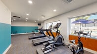 Located at 8560 W Peoria Ave in Peoria, Arizona, this fitness center offers a well-organized, single-floor exercise space designed to accommodate a full range of workout routines. The room features a vibrant two-tone color scheme, with a bold teal accent band running along the lower half of the walls, complemented by warm orange accents near the window area, all set against crisp white upper walls and a textured ceiling with recessed lighting.

The cardio section is equipped with multiple Star Trac treadmills, an elliptical cross-trainer, and a stationary spin bike, positioned along the windowed wall that offers direct views of an outdoor pool area, providing natural light and a pleasant visual backdrop during workouts. A wall-mounted flat-screen TV is installed in the free weights area for entertainment while exercising.

The strength training section includes a two-tier dumbbell rack with a full range of hex weights, a foam roller and medicine ball storage rack, a cable/pull-up station, and an adjustable workout bench. The carpeted flooring with linear patterning provides comfort underfoot throughout the space. A dedicated exit door and signage ensure safe and convenient access. Overall, the fitness center delivers a functional, thoughtfully appointed environment suited for both cardio and strength training.
