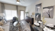 This single-floor space showcases a thoughtfully designed open-concept living and dining area that balances comfort with modern aesthetics. The living area features light-toned hardwood-style flooring that extends throughout, complemented by a plush area rug that anchors a seating arrangement consisting of a sleek accent chair and a round glass-top coffee table. A ceiling fan with a light fixture overhead provides both functionality and a subtle design element. Natural light filters generously into the space through multiple windows dressed with layered blinds and patterned curtains, creating a bright and welcoming atmosphere. A glass-paneled door provides access to an exterior space, further enhancing the sense of openness. The dining area is defined by a round glass dining table paired with upholstered chairs, positioned adjacent to the living space for a seamless flow. A dark wood console table along the wall serves as a stylish accent piece, topped with decorative objects and a table lamp. Two framed art pieces in silver frames add a curated, gallery-like touch to the neutral-toned walls. The overall ambiance is contemporary and inviting, with a cohesive neutral color palette punctuated by warm wood tones and metallic accents.