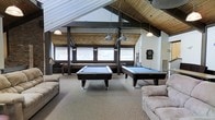 This single-floor recreational space is designed for leisure and socializing, highlighted by a dramatic vaulted ceiling with exposed dark wooden beams that add architectural character and a sense of openness. The angled roofline creates an expansive, airy feel throughout the room. Two full-sized pool tables with dark frames and blue felt surfaces serve as the centerpiece of the space, positioned beneath industrial-style pendant lights that provide focused illumination. A dartboard mounted on the wall offers an additional entertainment option. Comfortable, neutral-toned sofas line the perimeter of the room, creating casual seating areas for relaxation and socializing. Natural light filters in through a row of windows along one wall, framed by woven bamboo shades that contribute a warm, textured aesthetic. The neutral carpeting throughout ties the space together and provides a comfortable underfoot feel. Structural support columns and wood-paneled ceiling details further enhance the rustic yet functional design of the room, making this space well-suited for recreational use.