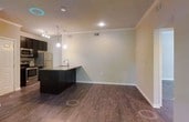 This single-floor space showcases a thoughtfully designed open-concept layout that seamlessly connects the kitchen and living areas. The kitchen is a standout feature, equipped with dark espresso cabinetry that provides ample storage, sleek granite countertops, and a full suite of stainless steel appliances including a refrigerator, range, and microwave. A central island with an integrated sink serves as both a functional workspace and a natural gathering point, accented by elegant pendant lighting overhead. A secondary track lighting fixture provides additional illumination to the cooking area. The open living space flows naturally from the kitchen, offering a generous and flexible area bathed in warm ambient light. Rich, dark hardwood-style flooring runs throughout, adding warmth and continuity to the entire space. Crown molding along the ceiling adds a refined architectural detail, while recessed lighting ensures the room remains bright and welcoming. Neutral wall tones create a versatile backdrop suitable for a variety of interior styles. The layout is efficient yet spacious, making excellent use of the single-floor footprint to deliver a comfortable and modern living environment.
