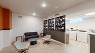 Located on Destination Drive in Broomfield, Colorado, this single-floor laundry facility combines practical functionality with thoughtful design. The space is divided into two distinct zones: a welcoming lounge area and a fully equipped laundry room.

The lounge area sets a polished, modern tone with light wood-look flooring, recessed lighting, and clean white walls accented by a dark feature wall. A set of three decorative framed laser-cut art panels with wood trim serves as a striking visual divider between the seating and laundry areas. Comfortable seating — including a navy sofa, swivel lounge chairs with woven frames, and a low round coffee table — creates an inviting space to relax while waiting.

The laundry room is efficiently laid out with a row of commercial-grade front-loading washers and dryers lining one wall, complemented by a built-in wooden folding bench with a mosaic tile accent along the base. A utility sink is also available for convenience. Natural light enters through a skylight and a small high window, keeping the space bright. The room is finished with the same light flooring for a cohesive aesthetic throughout.

Access between areas is provided via a glass-paneled door with a dark frame, and the facility is monitored by security cameras. Informational signage and a thermostat panel reflect the space's practical, resident-focused design.