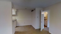 This single-floor space presents a straightforward and functional layout with a neutral color palette throughout. The main living area features wall-to-wall beige carpeting and smooth white walls accented by a continuous chair rail running along the perimeter, adding a subtle architectural detail. Overhead HVAC venting is integrated into the ceiling and walls, ensuring climate control throughout. A thermostat is conveniently mounted on the wall, and standard electrical outlets are positioned at regular intervals.

To the left, a partial view reveals a countertop surface with white cabinetry above, suggesting a kitchen or utility area nearby. Through an open doorway on the right, an adjoining room is visible, featuring hard flooring, light-toned cabinetry, and a warm overhead light, consistent with a kitchen or bathroom space. The transition between the carpeted living area and the hard-floored room provides a clear functional separation within the unit.

Overall, the space maintains a clean, minimal aesthetic with consistent white trim work, neutral tones, and practical finishes suited for comfortable everyday living.