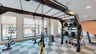 Located at 651 East 6th Street in Tempe, Arizona, this fitness center offers a generously sized, single-floor layout designed to accommodate a full range of workout activities. The space is thoughtfully organized to maximize usability, with distinct zones for cardio, strength training, and functional fitness.

The cardio area features a row of Echelon spin bikes and treadmills positioned along a wall of large windows, allowing natural light to flood in while providing views of the surrounding outdoor greenery and palm trees — a nod to the Arizona setting. Roller shades offer adjustable light control throughout the day.

The strength training section is outfitted with a selection of selectorized cable machines and plate-loaded equipment, complemented by large wall-mounted mirrors that visually expand the space and allow users to monitor their form. A functional training rig — prominently branded TRX — anchors the free weights zone, complete with kettlebells, weight plates, barbells, and a squat rack with a bench.

The flooring throughout features a distinctive checkered tile pattern in soft green and neutral tones, lending a modern, energetic aesthetic. Linear LED accent lighting runs along the ceiling, adding a sleek, contemporary touch. Decorative wall art, including an abstract mural in warm earth tones, adds a design-forward element to the space. The overall ambiance is bright, clean, and motivating.