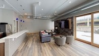 This contemporary single-floor space blends industrial and modern design elements to create a welcoming and functional environment. The open-plan layout is anchored by warm hardwood-style flooring that flows seamlessly throughout, complemented by high ceilings with exposed ductwork that add a distinctive industrial character.

The lounge area serves as the focal point of the space, featuring a tufted leather sectional sofa and coordinating accent chairs arranged around a central coffee table. A striking floor-to-ceiling mosaic tile accent wall houses both a wall-mounted flat-screen television and a sleek fireplace, creating an inviting gathering area. Expansive sliding glass doors and large windows flood the lounge with natural light and provide views of the exterior.

Adjacent to the lounge, the kitchen area is thoughtfully appointed with dark wood cabinetry, white subway tile backsplash, a stainless steel refrigerator, and a clean white countertop island. Distinctive blue pendant lights add a pop of color overhead. Multiple entry and exit points, including glass-paneled doors, ensure easy accessibility throughout the space. Recessed lighting and track lighting further enhance the bright, open ambiance, making this space well-suited for both social gatherings and everyday use.